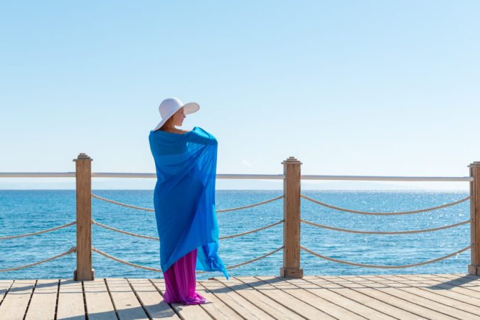 Femme sur une jetée en bois portant un grand chapeau et s'enveloppant dans un drapé bleu vif, parfait comme poncho pour la plage