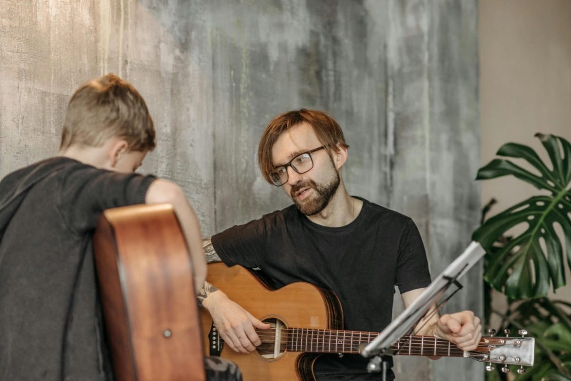 Un professeur de musique barbu donne un cours de guitar acoustique à un jeune élève dans un studio au style industriel
