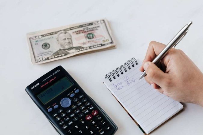 Une main écrit des notes financières à côté d'une calculatrice et de billets pour optimiser votre budget.